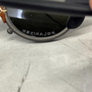 May include: Close-up of a pair of sunglasses with a dark gray frame and green lenses. The text "POLARIZED" is visible on the bottom of the frame. The temples have brown leather and cream stitching.