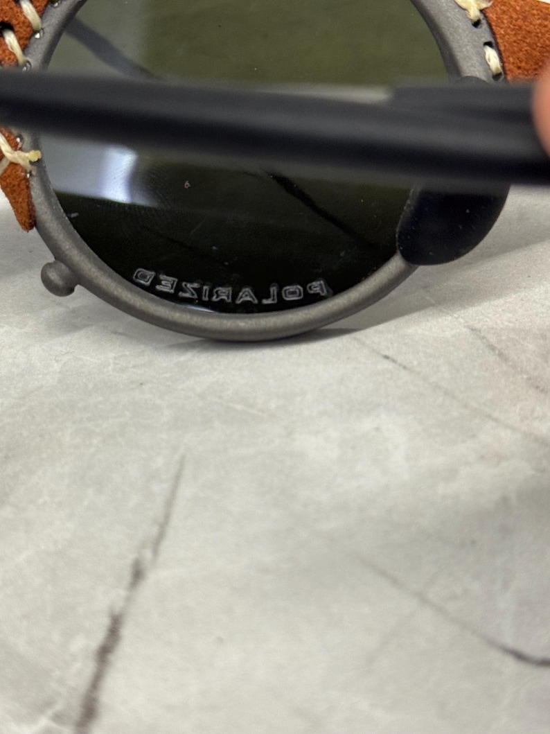 May include: Close-up of a pair of sunglasses with a dark gray frame and green lenses. The text "POLARIZED" is visible on the bottom of the frame. The temples have brown leather and cream stitching.