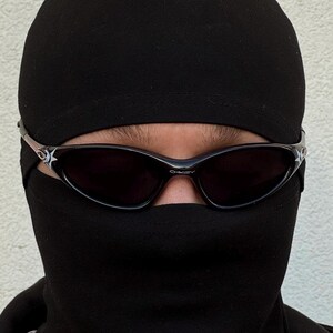 K&ouml;nnte beinhalten: Eine Person tr&auml;gt eine schwarze Sturmhaube und Sonnenbrille. Die Sonnenbrille hat dunkle Gl&auml;ser und einen silbernen Rahmen mit einem Sterndesign an den B&uuml;geln. Die Sturmhaube bedeckt Kopf und Hals.