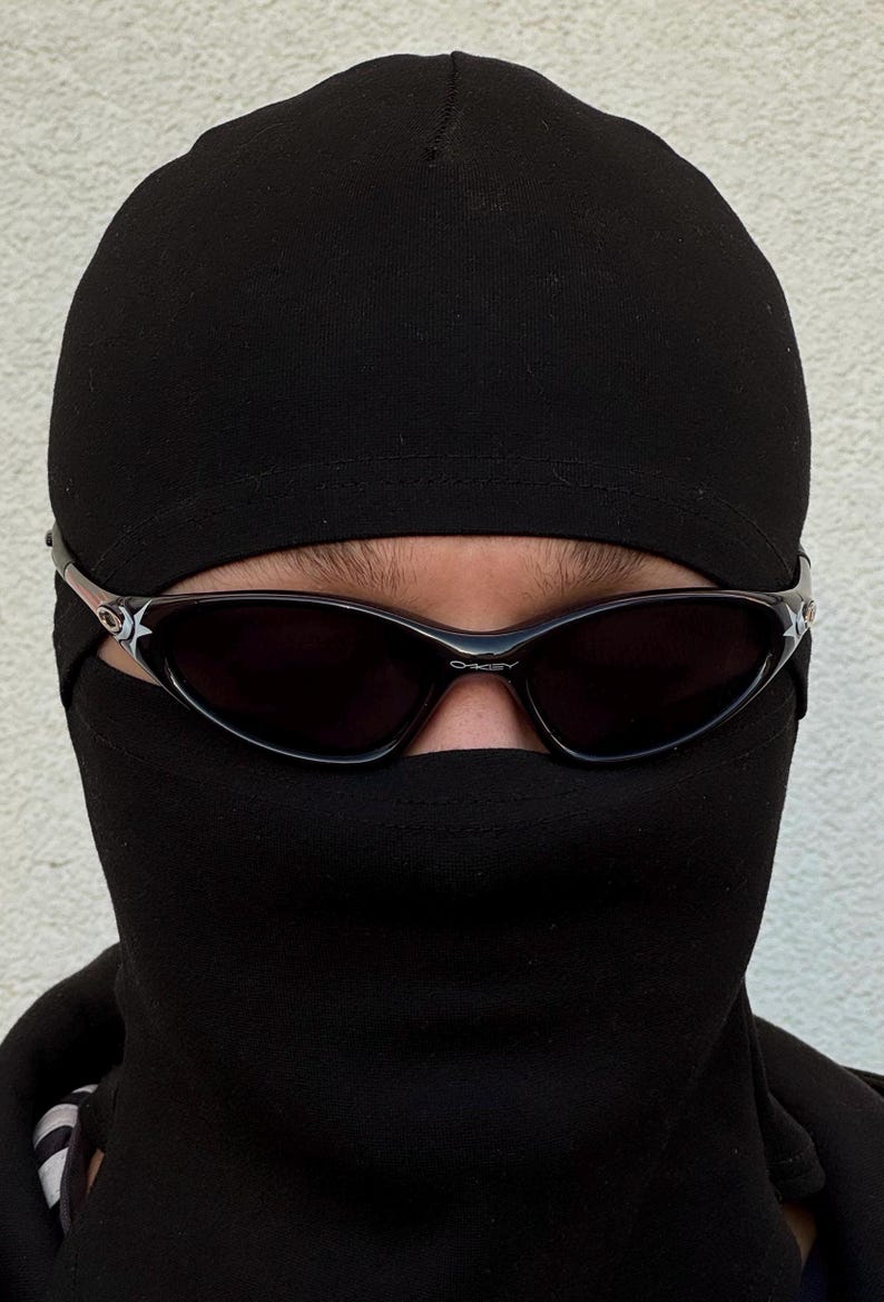 K&ouml;nnte beinhalten: Eine Person tr&auml;gt eine schwarze Sturmhaube und Sonnenbrille. Die Sonnenbrille hat dunkle Gl&auml;ser und einen silbernen Rahmen mit einem Sterndesign an den B&uuml;geln. Die Sturmhaube bedeckt Kopf und Hals.