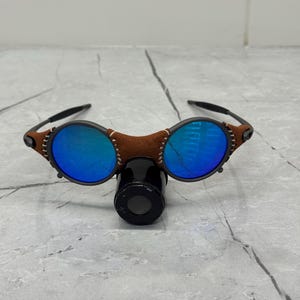 以下が含まれることがあります： 丸い青いレンズと茶色の革のブリッジが付いた黒いサングラス。 フレームは、目に見えるステッチと下の小さな黒いアタッチメントを備えたユニークなデザインです。