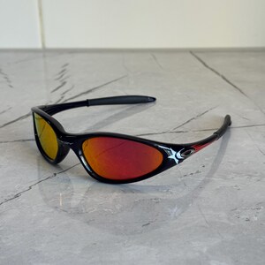 May include: Black sunglasses with red and yellow lenses. The sunglasses have a sleek, curved design with a black frame and red accents. The lenses reflect a gradient of red and yellow.