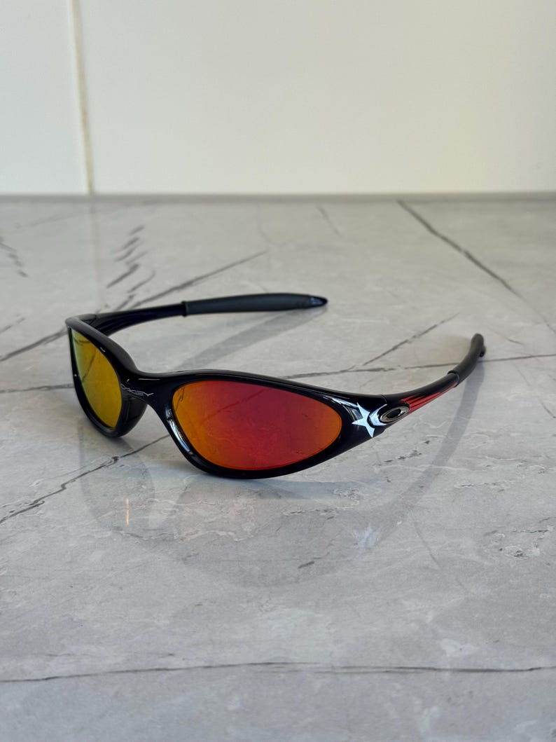 May include: Black sunglasses with red and yellow lenses. The sunglasses have a sleek, curved design with a black frame and red accents. The lenses reflect a gradient of red and yellow.