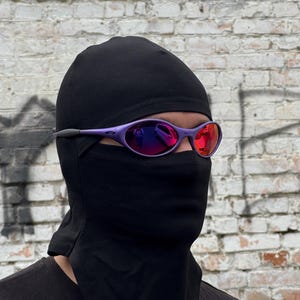 May include: A person wearing a black balaclava and purple sunglasses with red mirrored lenses. The sunglasses have a sporty, wraparound design. The background is a weathered brick wall.