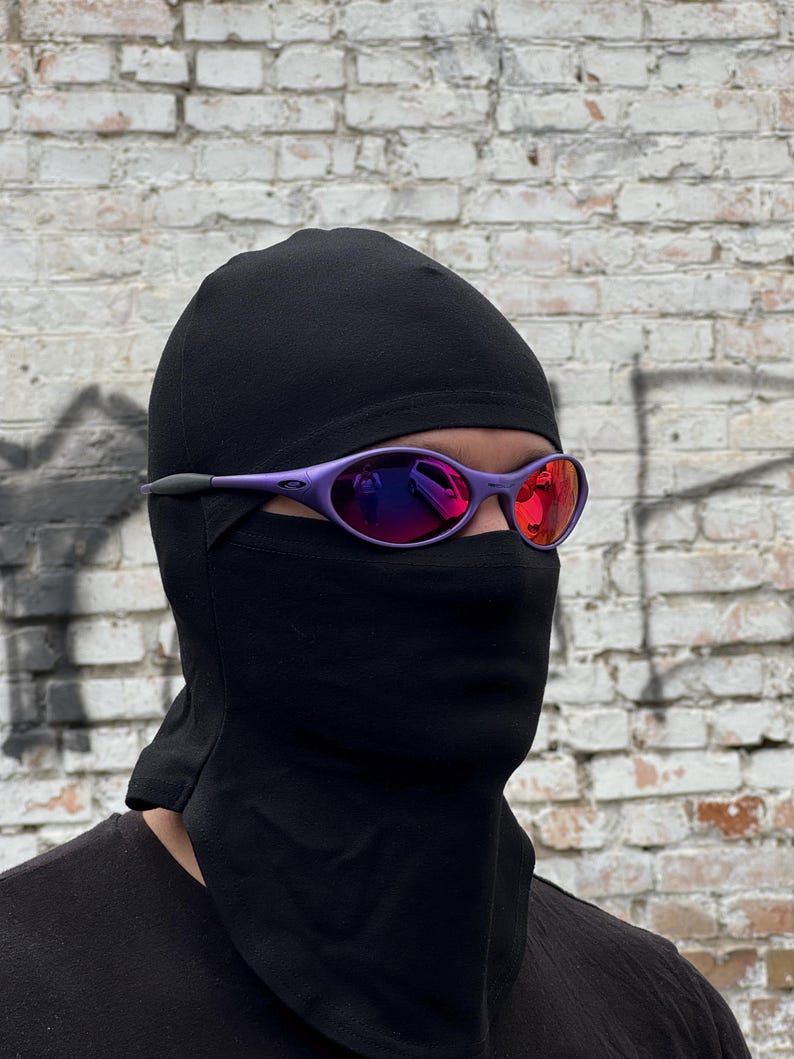 May include: A person wearing a black balaclava and purple sunglasses with red mirrored lenses. The sunglasses have a sporty, wraparound design. The background is a weathered brick wall.