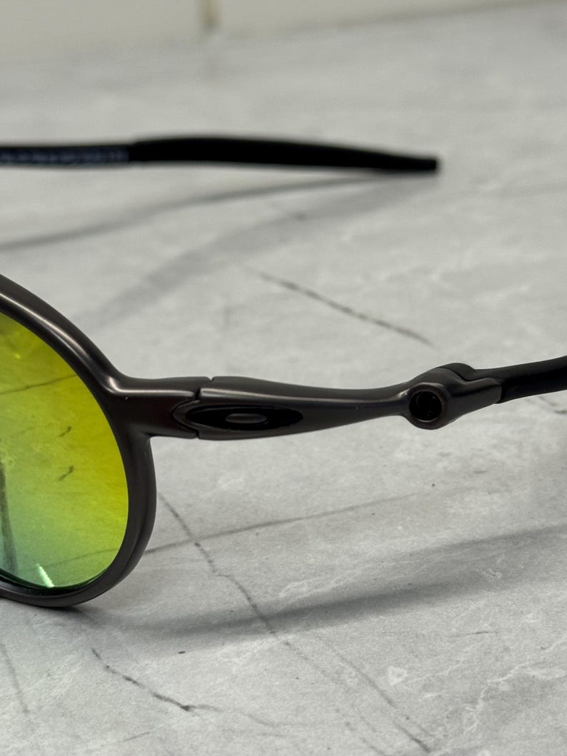 May include: Close-up of a pair of sunglasses with a dark gray frame and yellow-green lenses. The temples are black and extend to the ear. The lenses have a slight curve.