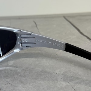 May include: Silver-framed sunglasses with black lenses. The text "MADE IN U.S.A." is printed on the silver arm. The sunglasses have a sleek, modern design.