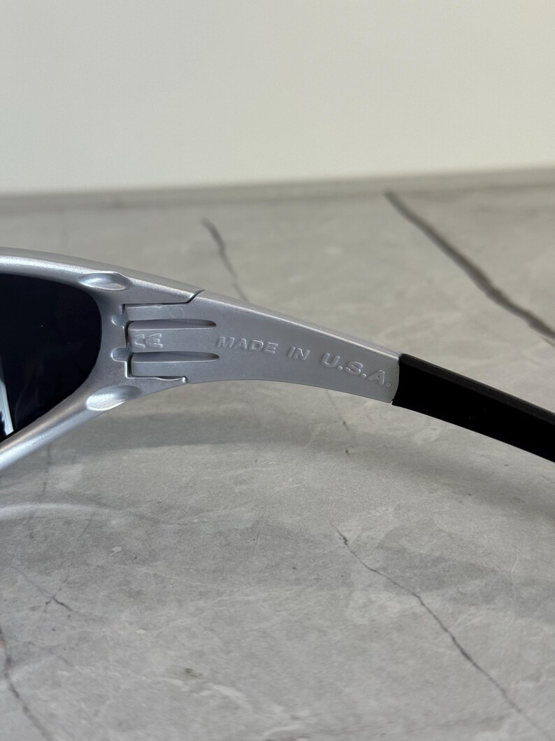 May include: Silver-framed sunglasses with black lenses. The text "MADE IN U.S.A." is printed on the silver arm. The sunglasses have a sleek, modern design.