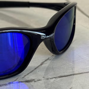 May include: Black sunglasses with blue lenses. The sunglasses have a sleek, modern design with a curved frame. The Oakley logo is visible on the side of the frame. The lenses are a vibrant blue color.