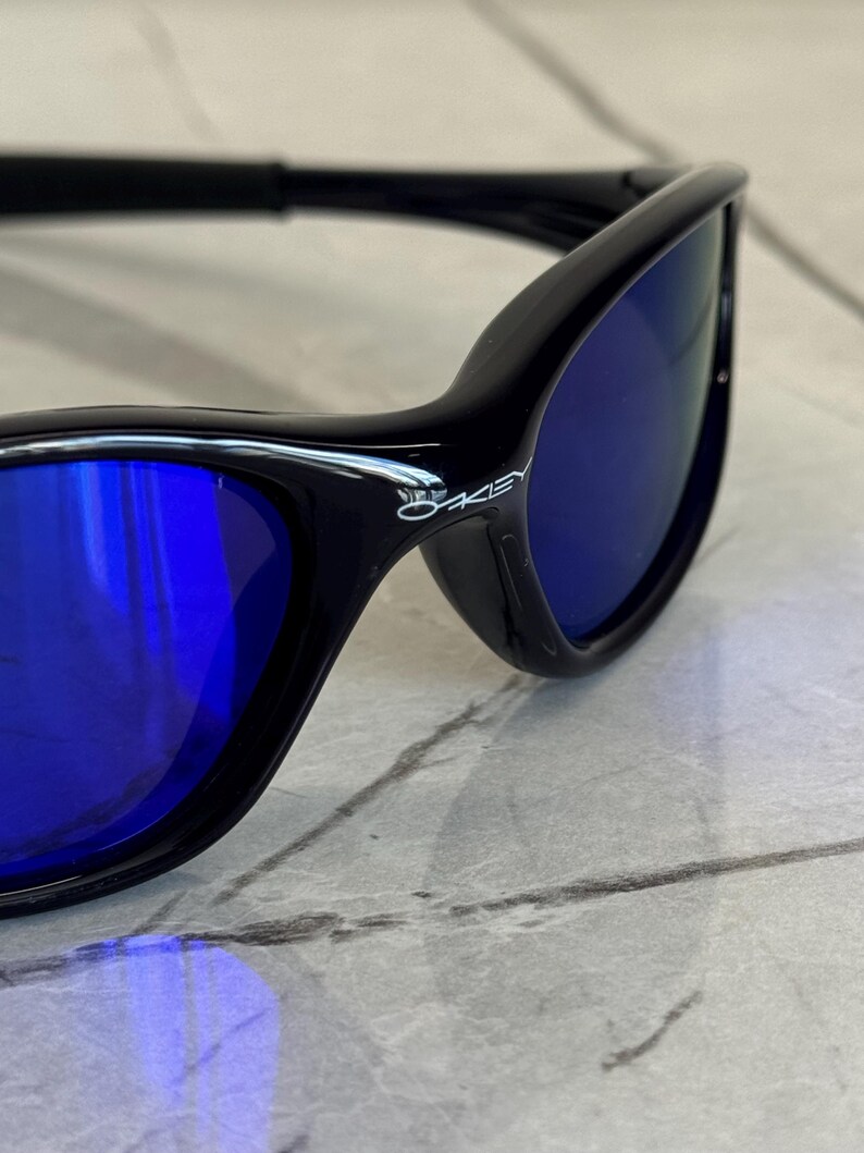 May include: Black sunglasses with blue lenses. The sunglasses have a sleek, modern design with a curved frame. The Oakley logo is visible on the side of the frame. The lenses are a vibrant blue color.