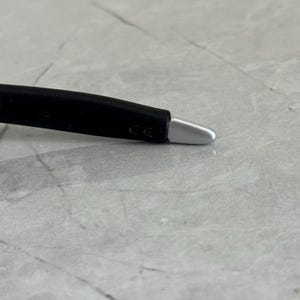 May include: Close-up of a black eyeglass arm with a silver tip. The arm has a curved design and the letters "CE" are visible. The background is a light grey, textured surface.