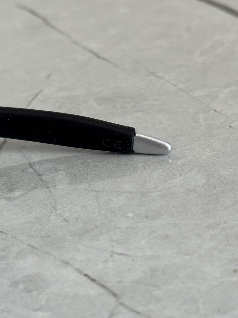 May include: Close-up of a black eyeglass arm with a silver tip. The arm has a curved design and the letters "CE" are visible. The background is a light grey, textured surface.