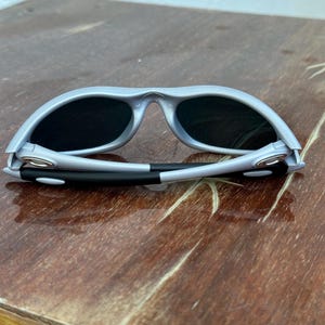 May include: Gray sunglasses with black lenses. The sunglasses have a curved frame and a black and white arm. The sunglasses are resting on a wooden surface.