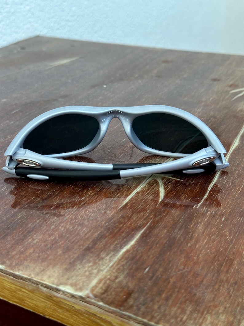 May include: Gray sunglasses with black lenses. The sunglasses have a curved frame and a black and white arm. The sunglasses are resting on a wooden surface.