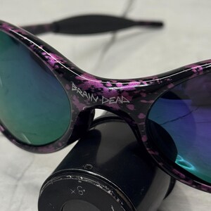 May include: Black sunglasses with a purple splatter design and mirrored lenses. The text "BRAIN DEAD" is printed on the frame. The sunglasses are resting on a black cylindrical object.