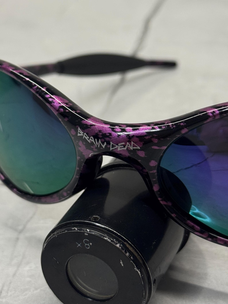 May include: Black sunglasses with a purple splatter design and mirrored lenses. The text "BRAIN DEAD" is printed on the frame. The sunglasses are resting on a black cylindrical object.