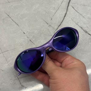 May include: Purple sunglasses with dark blue lenses. The sunglasses have a curved frame and are held by a hand. The lenses reflect a mix of blue and green light.