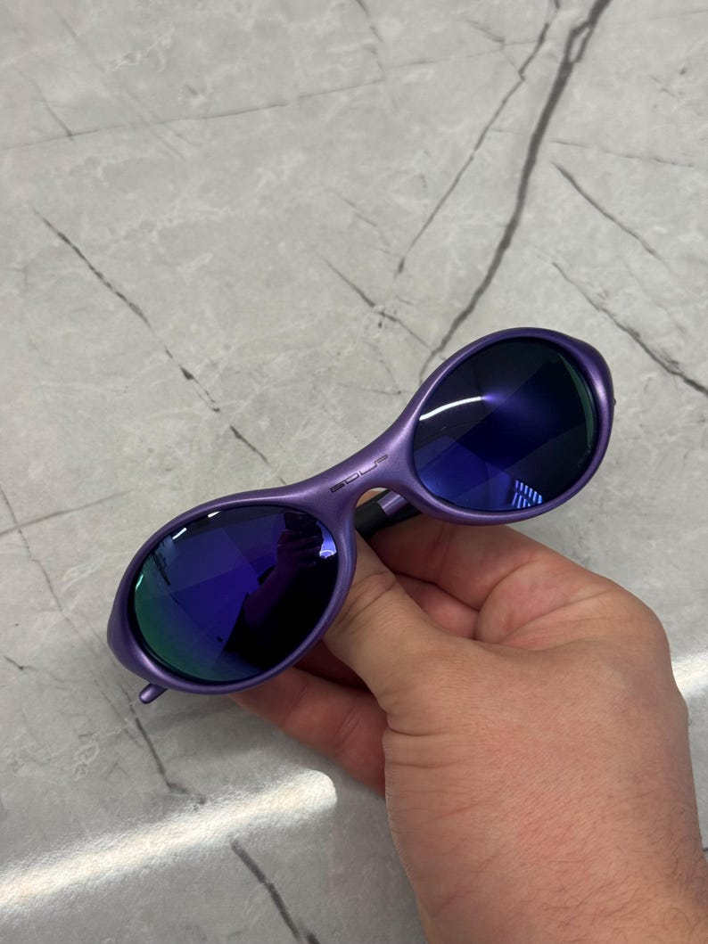May include: Purple sunglasses with dark blue lenses. The sunglasses have a curved frame and are held by a hand. The lenses reflect a mix of blue and green light.