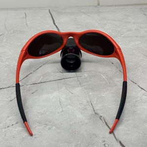 Op de afbeelding: Rode zonnebril met zwarte glazen en zwarte uiteinden van de pootjes. De zonnebril heeft een sportief, omsluitend ontwerp. Het montuur is felrood. De glazen zijn donker en bieden schaduw en bescherming tegen de zon.