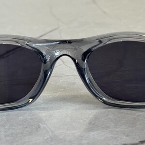 May include: Close-up of a pair of black sunglasses with a translucent gray frame. The lenses are dark, and the frame has a curved design. The sunglasses are likely for outdoor use.