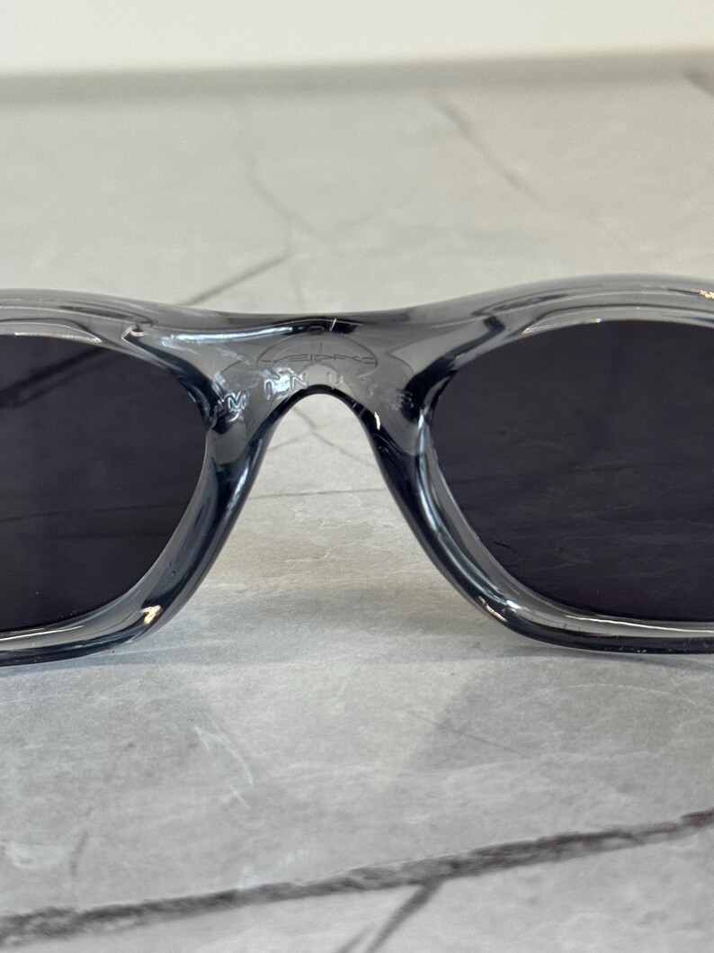 May include: Close-up of a pair of black sunglasses with a translucent gray frame. The lenses are dark, and the frame has a curved design. The sunglasses are likely for outdoor use.