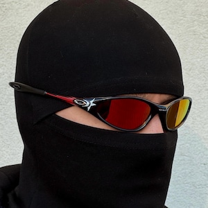 May include: Black sunglasses with red and black accents and mirrored lenses. The sunglasses have a star design on the side. The person is wearing a black balaclava.