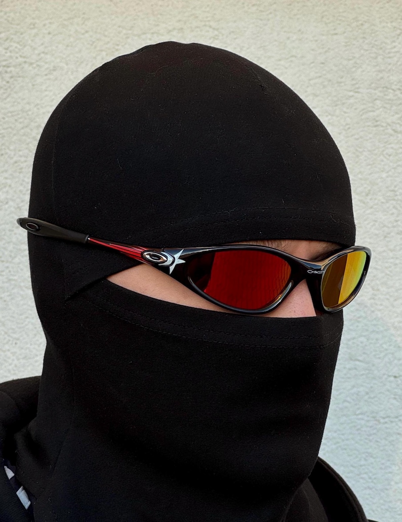May include: Black sunglasses with red and black accents and mirrored lenses. The sunglasses have a star design on the side. The person is wearing a black balaclava.