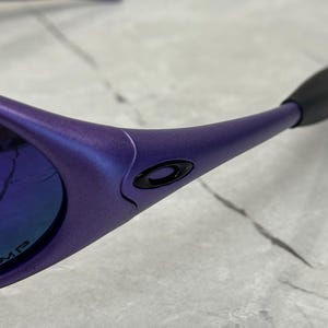 May include: Close-up of purple sunglasses with a black logo on the frame. The lenses have a blue and purple reflective coating. The arms of the sunglasses are a matching purple color.