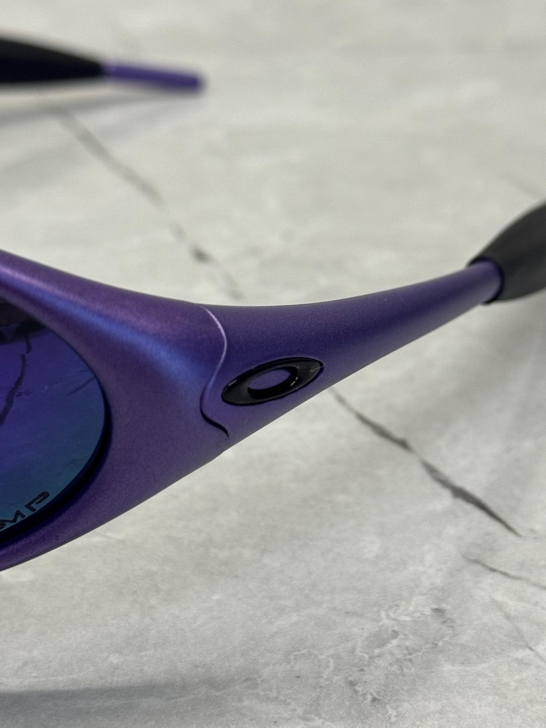 May include: Close-up of purple sunglasses with a black logo on the frame. The lenses have a blue and purple reflective coating. The arms of the sunglasses are a matching purple color.