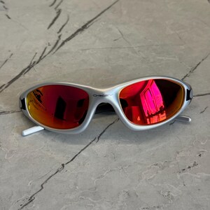 May include: Silver-framed sunglasses with red mirrored lenses. The sunglasses have a sleek, modern design with a curved frame and white arms. The Oakley logo is visible on the frame. The lenses reflect a bright pink hue.