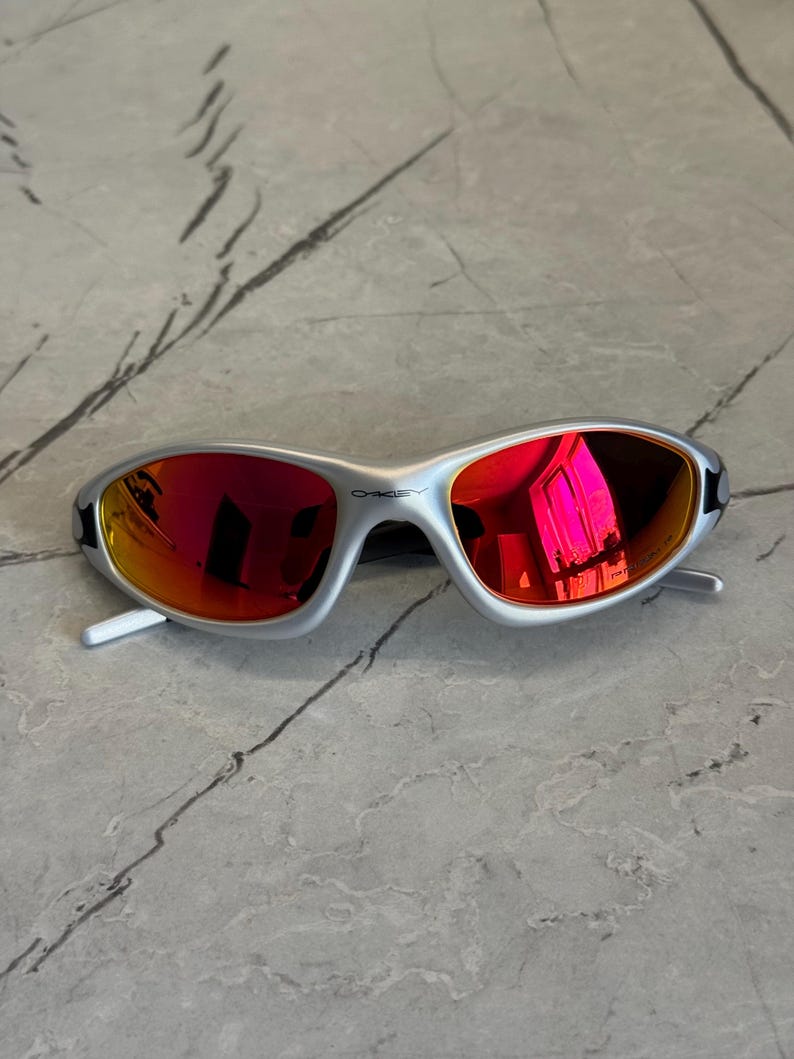 May include: Silver-framed sunglasses with red mirrored lenses. The sunglasses have a sleek, modern design with a curved frame and white arms. The Oakley logo is visible on the frame. The lenses reflect a bright pink hue.