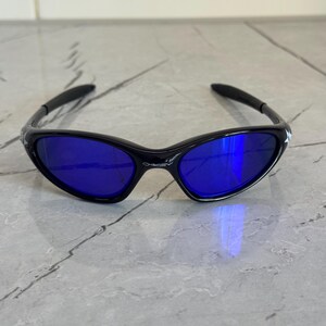 May include: Black sunglasses with blue lenses. The sunglasses have a sleek, curved frame design. The lenses are a vibrant blue color, reflecting light. The arms of the sunglasses are black and curve around the ears.