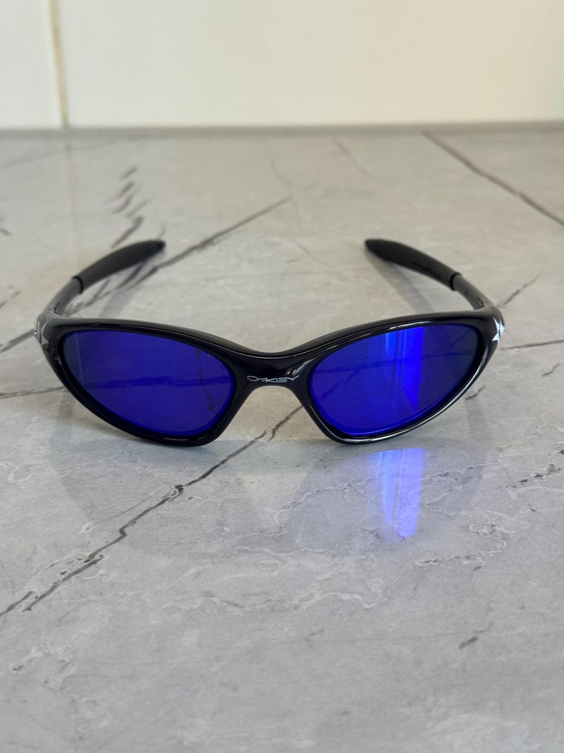 May include: Black sunglasses with blue lenses. The sunglasses have a sleek, curved frame design. The lenses are a vibrant blue color, reflecting light. The arms of the sunglasses are black and curve around the ears.