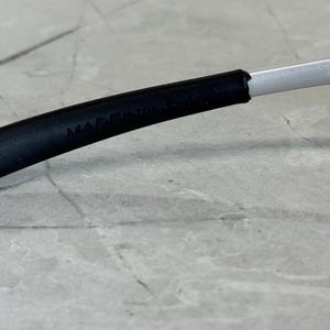 May include: Close-up of a black eyeglass frame with the text "MADE IN USA" visible. The frame transitions to a silver arm. The background is a light grey, textured surface.