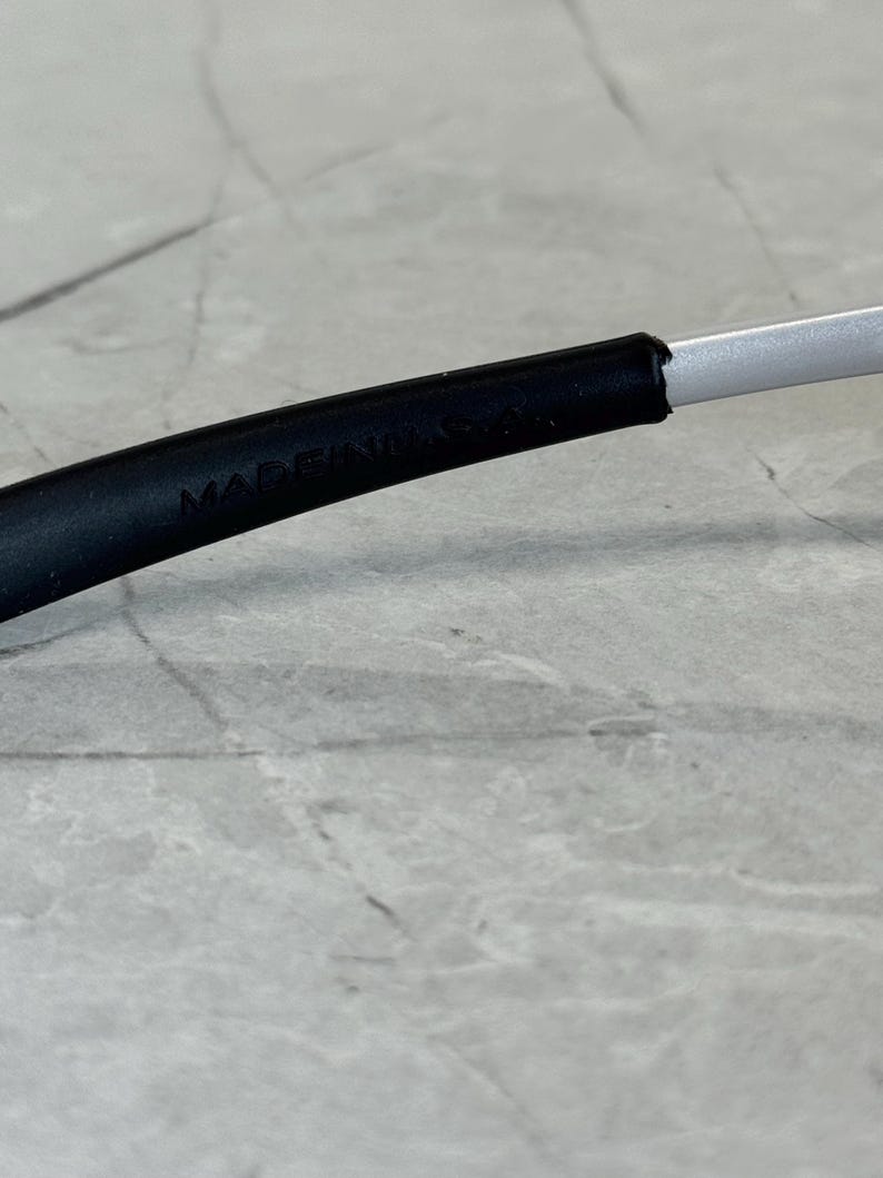May include: Close-up of a black eyeglass frame with the text "MADE IN USA" visible. The frame transitions to a silver arm. The background is a light grey, textured surface.