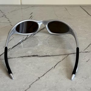 May include: Silver sunglasses with dark lenses. The sunglasses have a streamlined design with a curved frame and black temple tips. The lenses are oval-shaped, contrasting with the silver frame. A modern eyewear accessory.