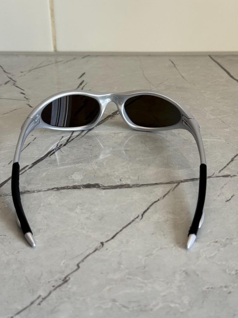 May include: Silver sunglasses with dark lenses. The sunglasses have a streamlined design with a curved frame and black temple tips. The lenses are oval-shaped, contrasting with the silver frame. A modern eyewear accessory.