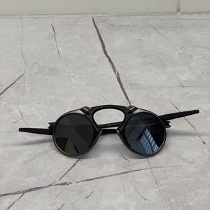 May include: Black sunglasses with circular lenses and a distinctive bridge. The frames are black with silver detailing. The arms extend outwards from the sides.