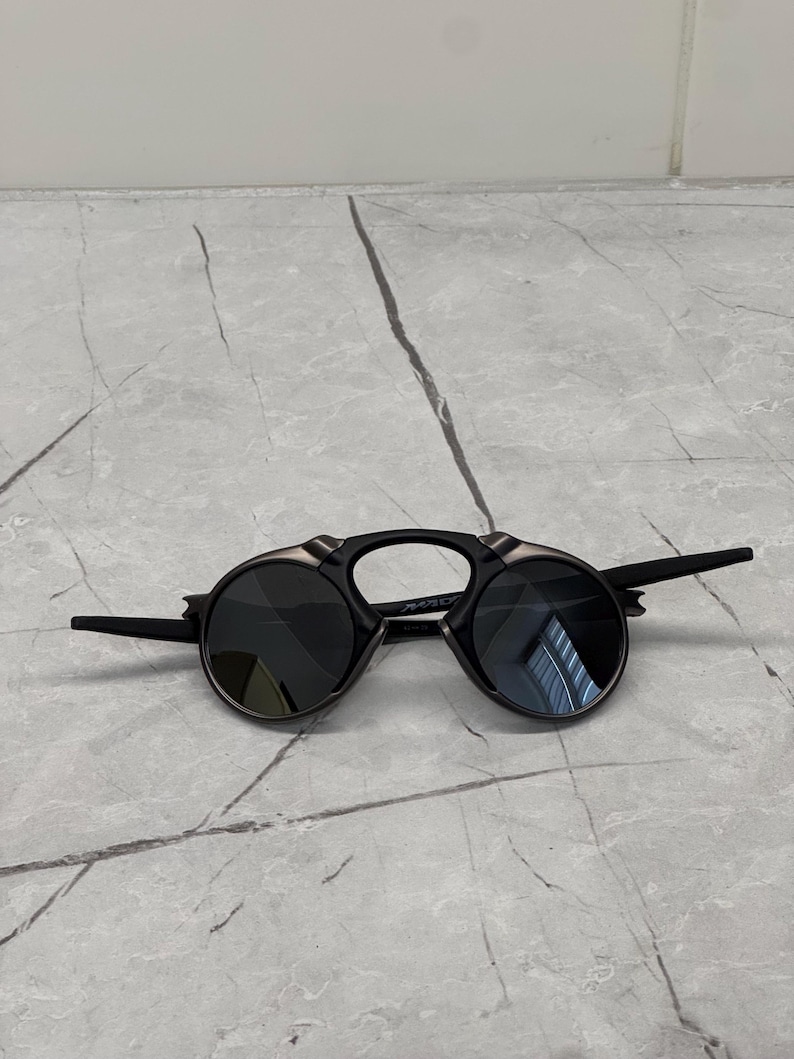 May include: Black sunglasses with circular lenses and a distinctive bridge. The frames are black with silver detailing. The arms extend outwards from the sides.
