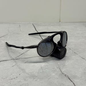 May include: Black sunglasses with a distinctive design, featuring circular lenses and a cylindrical component. The sunglasses have a sleek, modern aesthetic, with a matte finish and dark lenses. The arms are thin and curved.