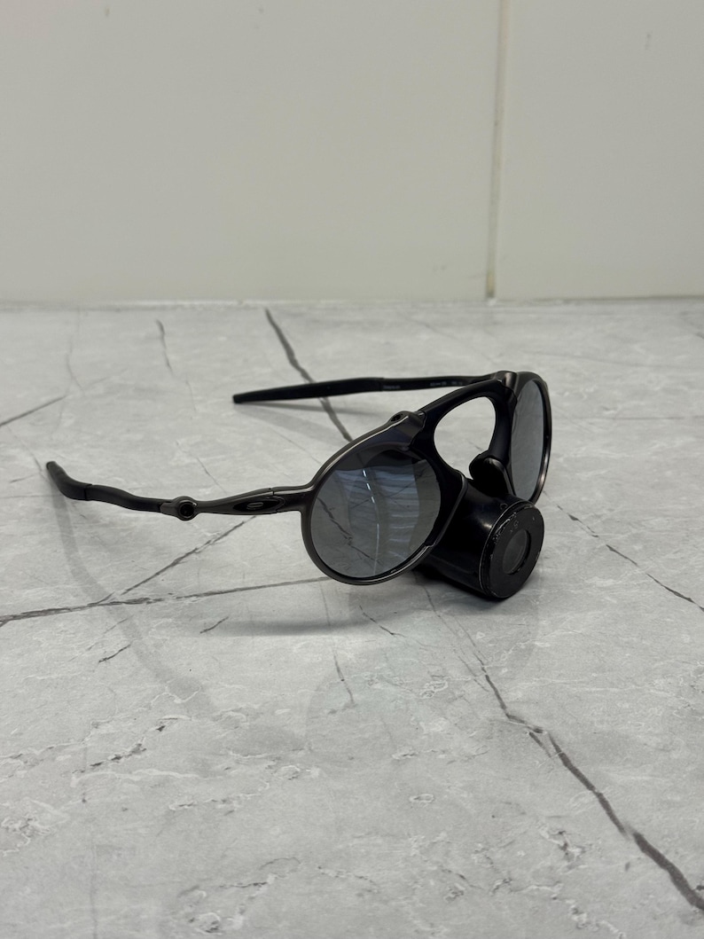 May include: Black sunglasses with a distinctive design, featuring circular lenses and a cylindrical component. The sunglasses have a sleek, modern aesthetic, with a matte finish and dark lenses. The arms are thin and curved.