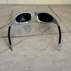 May include: Silver-framed sunglasses with black lenses. The sunglasses have a sleek, modern design with a curved frame and black temples. The lenses are dark, providing protection from the sun. The sunglasses are resting on a marble surface.