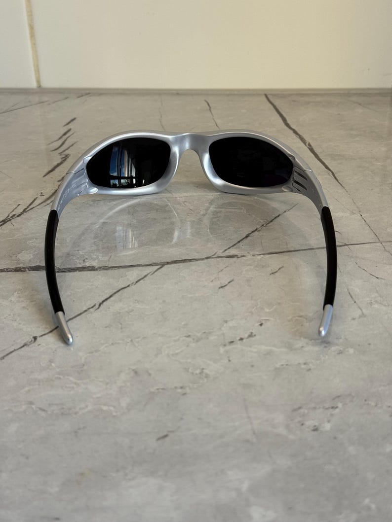 May include: Silver-framed sunglasses with black lenses. The sunglasses have a sleek, modern design with a curved frame and black temples. The lenses are dark, providing protection from the sun. The sunglasses are resting on a marble surface.