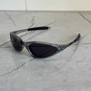May include: Clear gray sunglasses with dark lenses. The sunglasses have a sleek, sporty design with a curved frame and black temple tips. The lenses are oval-shaped and provide full coverage.