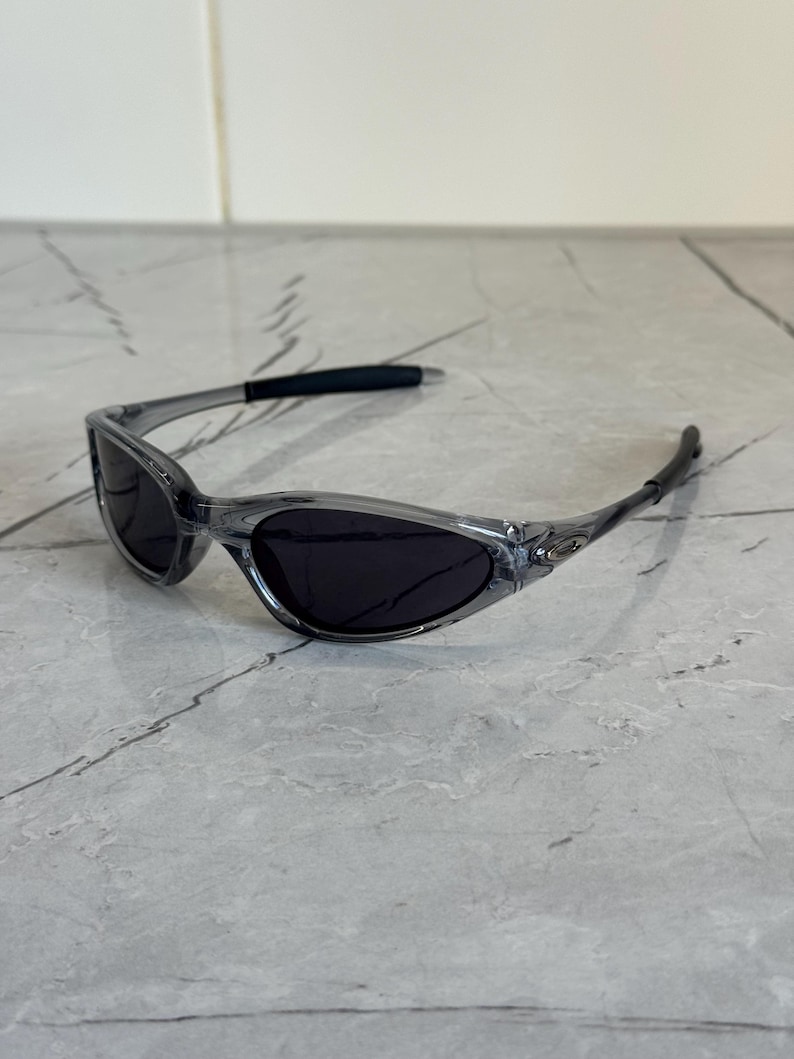 May include: Clear gray sunglasses with dark lenses. The sunglasses have a sleek, sporty design with a curved frame and black temple tips. The lenses are oval-shaped and provide full coverage.