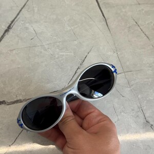 Puede incluir: Gafas de sol plateadas con lentes ovaladas y tinte oscuro. Las patillas tienen detalles azules. Las gafas de sol se sostienen sobre una superficie de m&aacute;rmol gris.