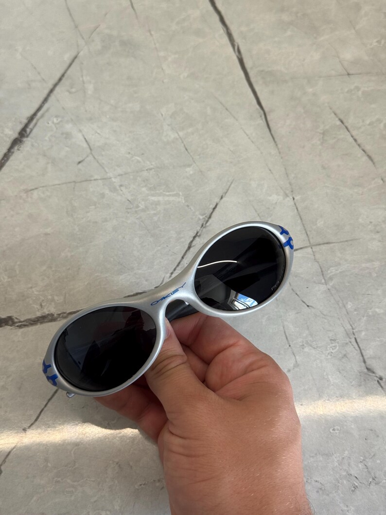 Puede incluir: Gafas de sol plateadas con lentes ovaladas y tinte oscuro. Las patillas tienen detalles azules. Las gafas de sol se sostienen sobre una superficie de m&aacute;rmol gris.