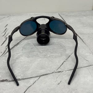 Pu&ograve; includere: Occhiali da sole con montatura nera, lenti blu e un accessorio nero. Gli occhiali da sole hanno un design elegante e moderno. Le lenti sono di un blu vibrante. La montatura &egrave; nera opaca.
