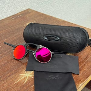May include: Black rimless sunglasses with red and pink lenses are displayed with a black carrying case and two black cleaning cloths. The case and cloths have the brand name "Oakley" printed on them. The sunglasses are on a wooden surface.