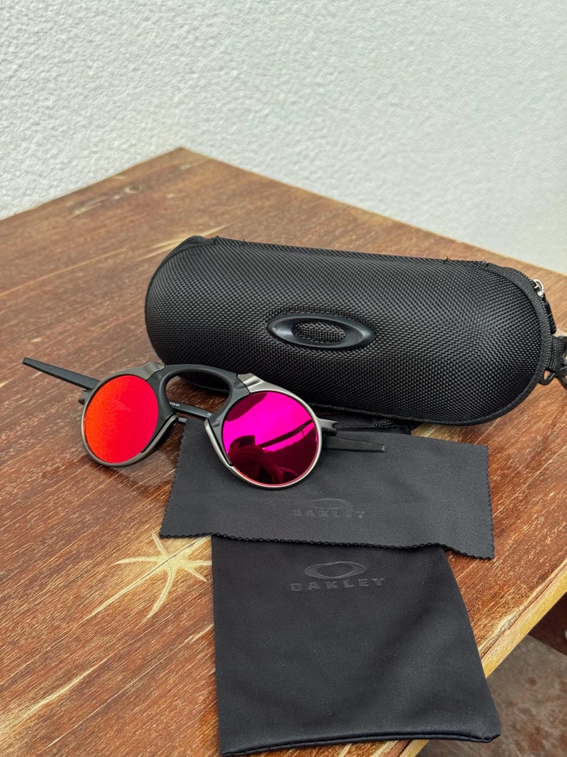 May include: Black rimless sunglasses with red and pink lenses are displayed with a black carrying case and two black cleaning cloths. The case and cloths have the brand name "Oakley" printed on them. The sunglasses are on a wooden surface.